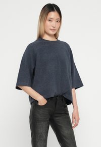 Mörkgrå oversized stickad tröja med korta ärmar och rundad halsringning, i kombination med svarta strukturerade byxor. Enkel, avslappnad design.