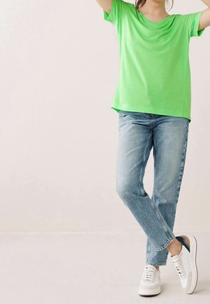 Persoon in een felgroen shirt met korte mouwen, lichte spijkerbroek en witte sneakers, staand met gekruiste benen tegen een effen achtergrond.