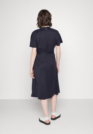 Vrouw met schouderlang bruin haar, gekleed in een zwarte midi-jurk en witte sandalen, staand met de rug naar de camera tegen een plain witte achtergrond.