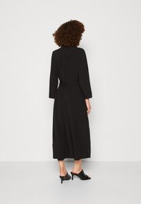 Abito midi nero con maniche lunghe, vita stretta e tessuto fluido. Presenta un sottile dettaglio plissettato sul retro. Indossato con mule neri con tacco.