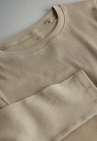 Bēšā ribotā garām piedurknēm krekls ar noapaļotu kakla izgriezumu un kabatu priekšpusē, ar mīkstu tekstūru un smalkiem šuvju elementiem.