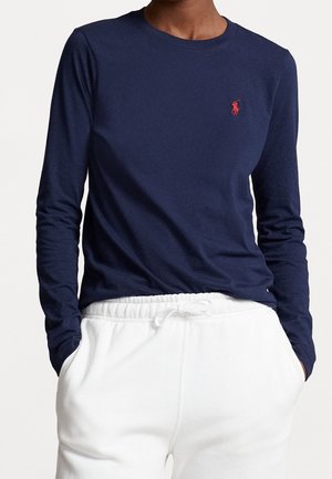 Personne portant une chemise bleu marine à manches longues avec un logo rouge et un pantalon blanc à cordon, mains dans les poches, torse visible.