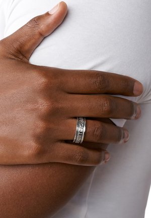 Anillo de banda de plata con grabado, que presenta un acabado suave y pulido. La mano del usuario descansa sobre una superficie de tela blanca.