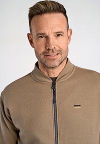 Giacca zip-up marrone chiaro con colletto alto. Presenta una cerniera blu contrastante e un piccolo logo metallico sul petto. Texture morbida.