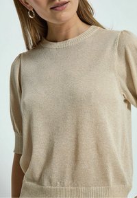 Maglione beige lavorato a maglia con maniche corte a sbuffo, scollo e orlo a coste. Texture morbida con sottili accenti scintillanti su tutta la superficie.