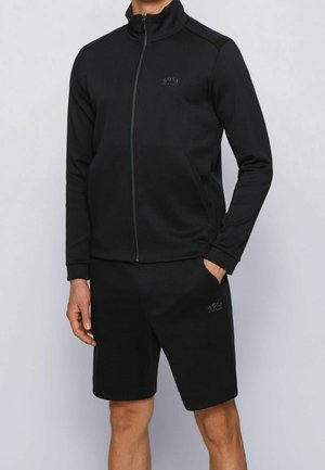 Mann trägt eine schwarze Jacke mit Reißverschluss und dazu passende schwarze Shorts, steht mit einer Hand in der Tasche vor einem einfarbigen hellen Hintergrund.