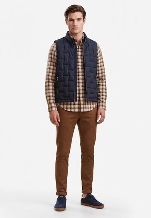 Joven con chaleco acolchado azul marino sobre una camisa de cuadros beige, pantalones marrones y zapatillas azul marino, de pie sobre un fondo blanco.