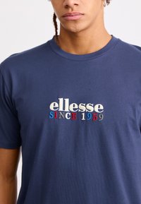 Navy blue cotton t-shirt featuring the embroidered text "ellesse SINCE 1959" in white, red, and blue. Classic crew neck design.