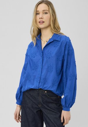 Chemise en coton bleu avec broderie florale, boutonnage devant, col ouvert et manches longues avec poignets élastiques. Portez avec un pantalon en denim foncé.