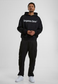 Hoodie noir avec le logo "visages oubliés" en blanc, associé à un pantalon cargo noir avec plusieurs poches et des baskets blanches.