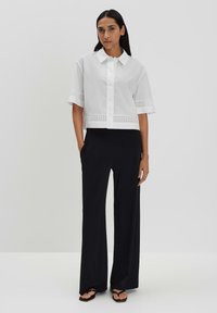 White cropped shirt with button closure, short sleeves, and eyelet trim detail, paired with black wide-leg pants and black sandals.