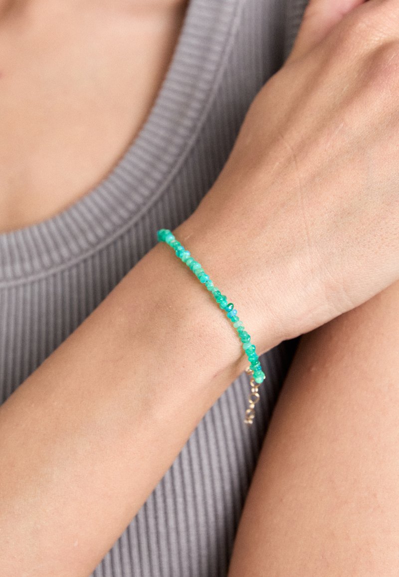 Pulsera de piedra turquesa con pequeñas cuentas irregulares y un broche de oro, llevada en una muñeca sobre un fondo gris texturizado.