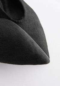 Par svarta skor med spetsiga tår, med en slät, matt yta och subtil textur synlig på ytan.