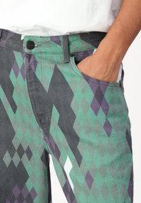 Pantalons à motif argyle vert, violet et gris avec une coupe droite, dotés d'une fermeture à bouton et d'une poche latérale. Détails de tissu texturé visibles.