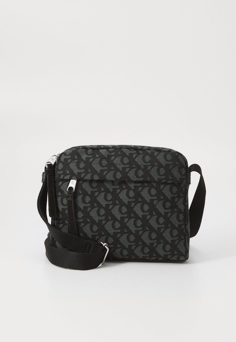 Czarna torba typu crossbody z geometrycznym wzorem, podwójnym zamkiem błyskawicznym, regulowanym paskiem i gładką fakturą. Posiada akcenty z srebrnego metalu.