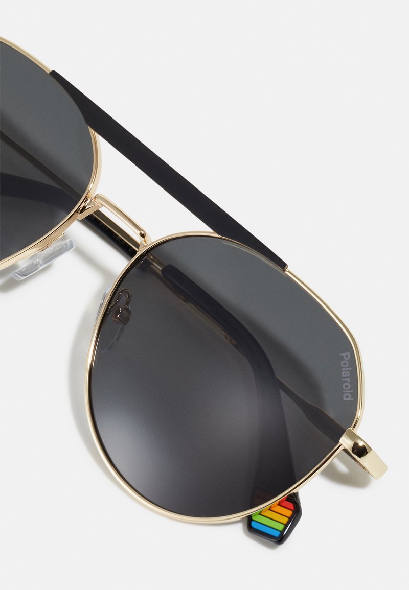 Gafas de sol aviador con montura dorada, lentes oscuros, patillas negras y un pequeño detalle de colores del arcoíris cerca de la bisagra sobre un fondo blanco.