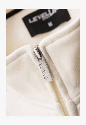 Gros plan d'une fermeture éclair blanche avec un curseur argenté gravé "OMBRE" sur un vêtement en tissu doux montrant une étiquette noire avec "LEVEL UP M".