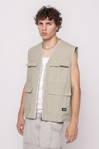 Light beige sleeveless vest with two chest pockets, front zipper, and logo tag. Fabric has a subtle texture, paired with gray denim pants.