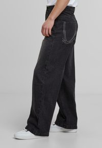 Jeans anchos de color negro hechos de mezclilla, con costuras en contraste blancas, bolsillos delanteros y traseros, y un ajuste relajado. Combinados con zapatillas blancas.