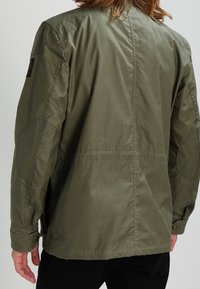 Olivově zelená bunda volného střihu, zdobená vzorovanými nášivkami na ramenou, s knoflíkovými manžetami a hladkým, mírně lesklým povrchem.