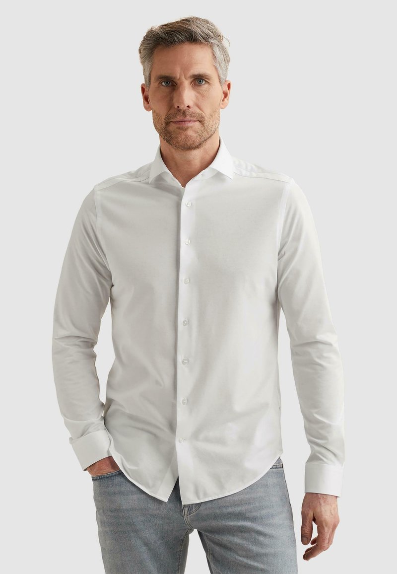Homme aux cheveux gris et à la barbe portant une chemise blanche ajustée boutonnée et un jean bleu clair, debout devant un fond uni.