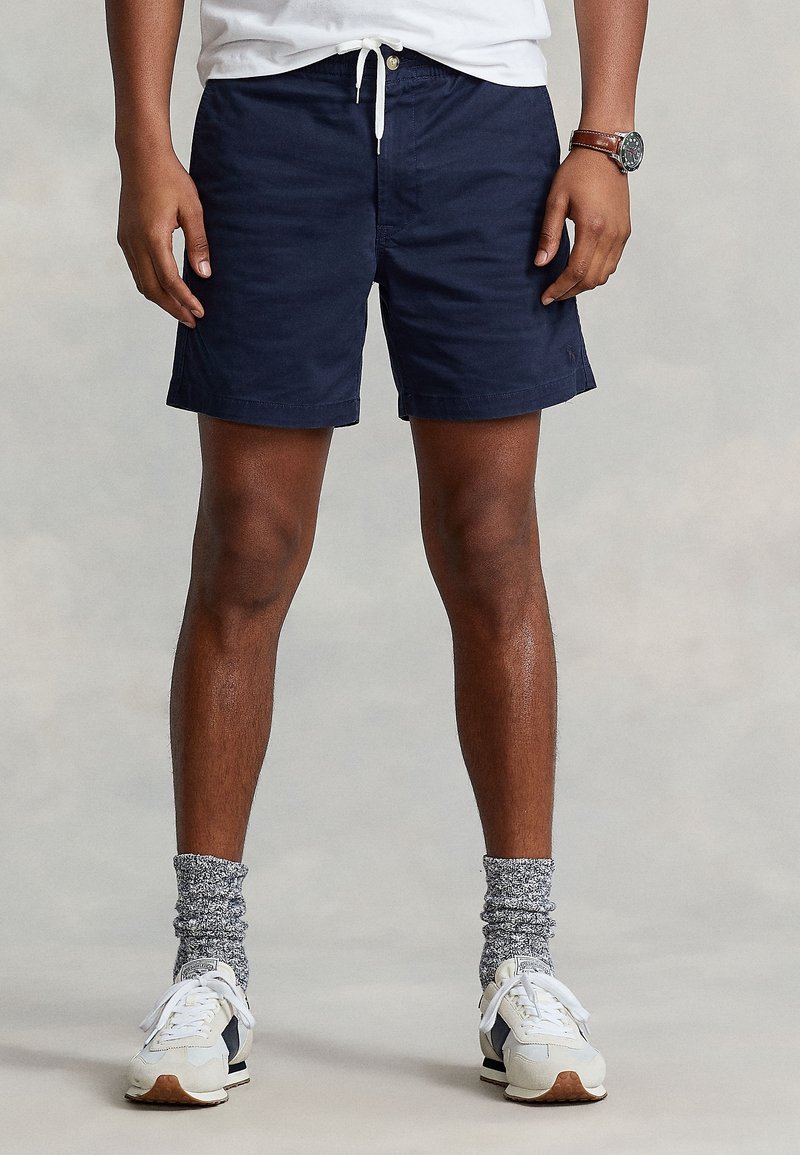 Homme portant un short à cordon de serrage bleu marine, des chaussettes gris chiné, des baskets blanches, un t-shirt blanc et une montre-bracelet, debout devant un fond neutre.