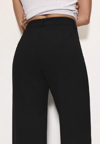 Pantalon noir à jambes larges en tissu lisse, doté d'une taille ajustée et de poches arrière discrètes, créant une silhouette épurée.