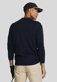 Navy lightweight knit sweater with long sleeves, ribbed cuffs, and hem. Worn with light-colored trousers and black gloves.