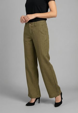 Donna che indossa pantaloni larghi color verde oliva, top nero aderente a maniche corte e tacchi neri con punta a punta, in piedi su sfondo grigio.