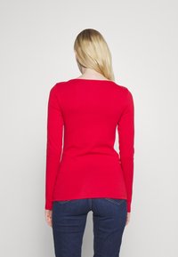 Maglietta rossa a maniche lunghe con scollo rotondo, realizzata in tessuto liscio. Design aderente, leggermente più lunga sul retro, abbinata a jeans in denim scuri.