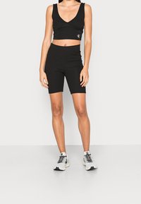 Kvinna i svart kort topp och svarta högmidjade cykelshorts som står mot en enkel ljus bakgrund.