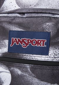 Jansport ruksak sa teksturiranim crno-belim materijalom, vidljivim zatvaračem i plavom oznakom sa crvenim brendiranjem.