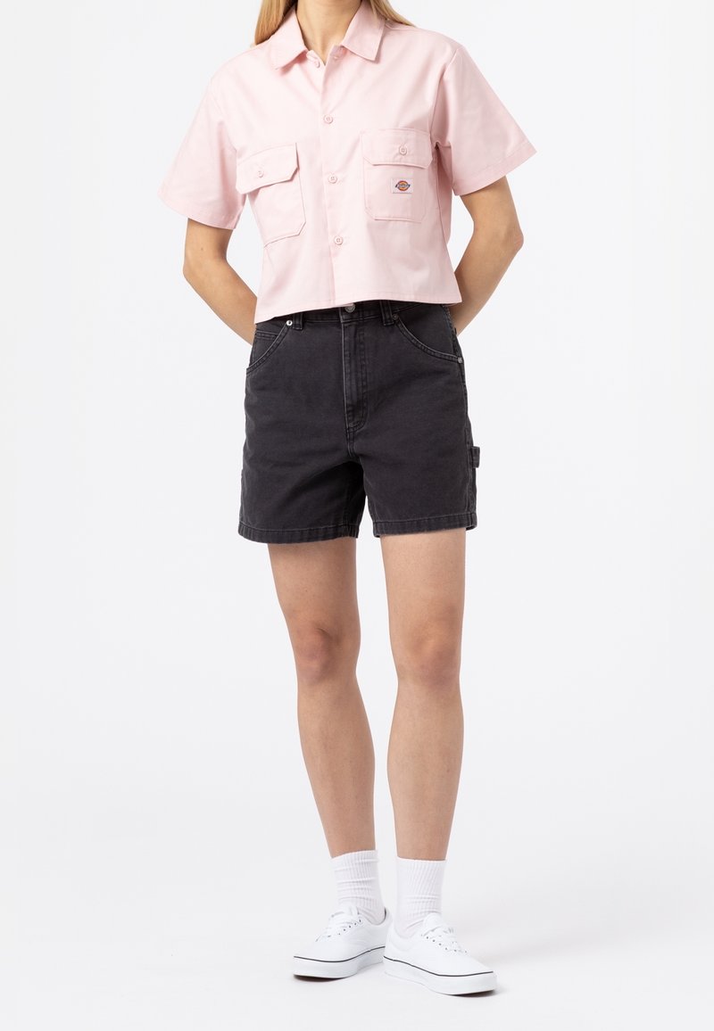 Femme portant une chemise rose clair à manches courtes avec des poches poitrine, un short en jean noir, des chaussettes blanches et des baskets blanches.