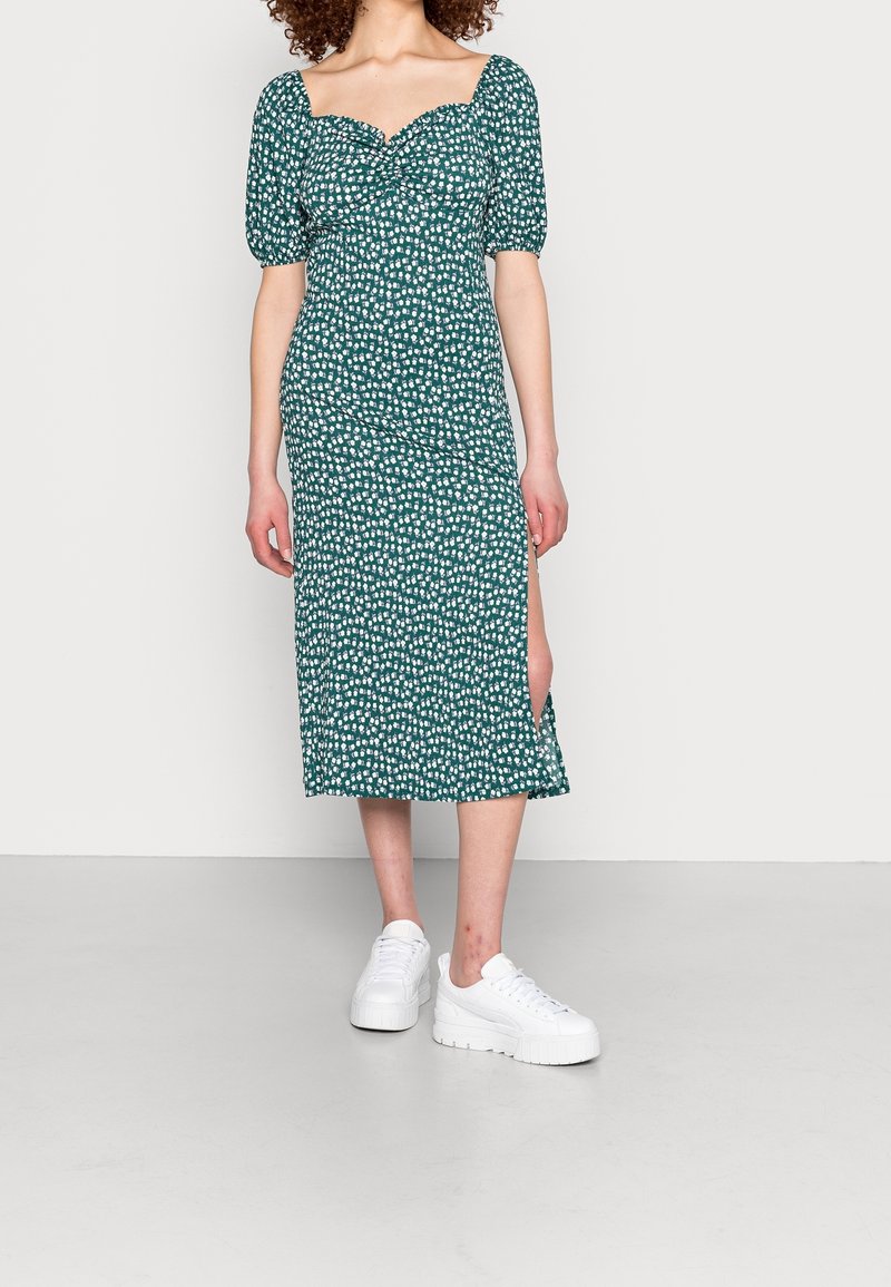 Femme portant une robe midi verte à imprimé floral blanc, manches bouffantes, fente latérale, et baskets blanches épaisses, debout devant un fond uni.