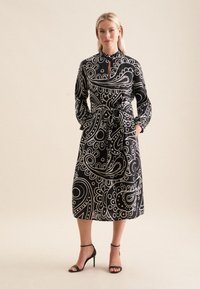 Schwarzes und weißes Kleid mit Paisleymuster, langen Ärmeln, einem geknöpften Kragen und einem gebundenen Taillenbereich. Glatte Stoffqualität mit mittellanger Schnittführung.
