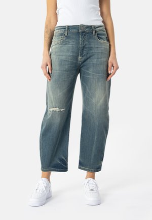 Vrouw draagt losse, vervaagde blauwe spijkerbroek met een klein scheurtje op de linker dij en witte sneakers, staand tegen een eenvoudige achtergrond.