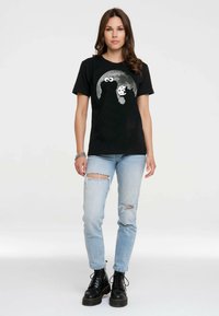 Schwarzes Baumwoll-T-Shirt mit einem grafischen Design eines Charakters mit großen Augen und einem Keks. Kombiniert mit hellblauen, zerrissenen Jeans und schwarzen Stiefeln.