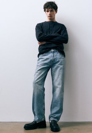 Schwarzes Henley-Shirt mit langen Ärmeln und Knöpfen vorne, kombiniert mit hellblauen, locker sitzenden Jeans und schwarzen Schuhen mit klobiger Sohle. Durchgehend glatte Texturen.