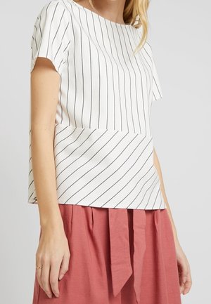 Vrouw draagt een wit kortärmelig blouse met dunne verticale en diagonale zwarte strepen, gecombineerd met een hooggetailleerde roze rok met een stoffen riem.