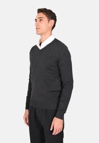 Pullover grigio scuro a maglia con scollo a V, maniche lunghe e polsini a coste, indossato sopra una camicia bianca con colletto.