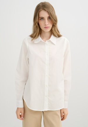 Joven mujer con cabello rubio hasta los hombros, que lleva una camisa blanca abotonada y pantalones beige frente a un fondo liso.