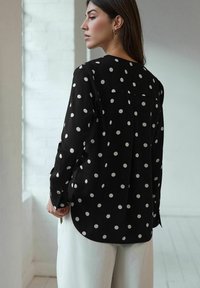 Schwarze Bluse mit weißen Polka-Dots, langen Ärmeln, abgerundetem Saum und einem Rückenteil mit Yoke-Details. Aus glattem Stoff gefertigt, im lockeren Schnitt gestaltet.