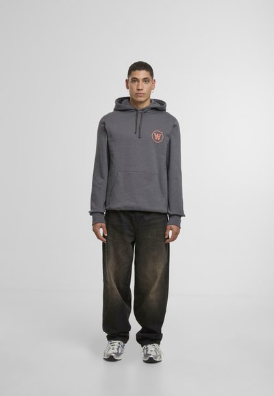 Grå hettegenser med et sirkulært oransje logo på brystet, sammen med løstsittende mørke jeans og grå, hvite og blå joggesko.