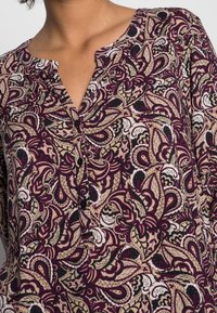 Camicia con motivo paisley viola e beige, con colletto a fessura e bottoni neri. Presenta vortici intricati e design floreali su un tessuto morbido.