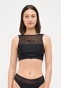Svart crop top med en transparent mesh front, brodert logo og en figursydd design. Materialet har en glatt tekstur med en cropped cut.