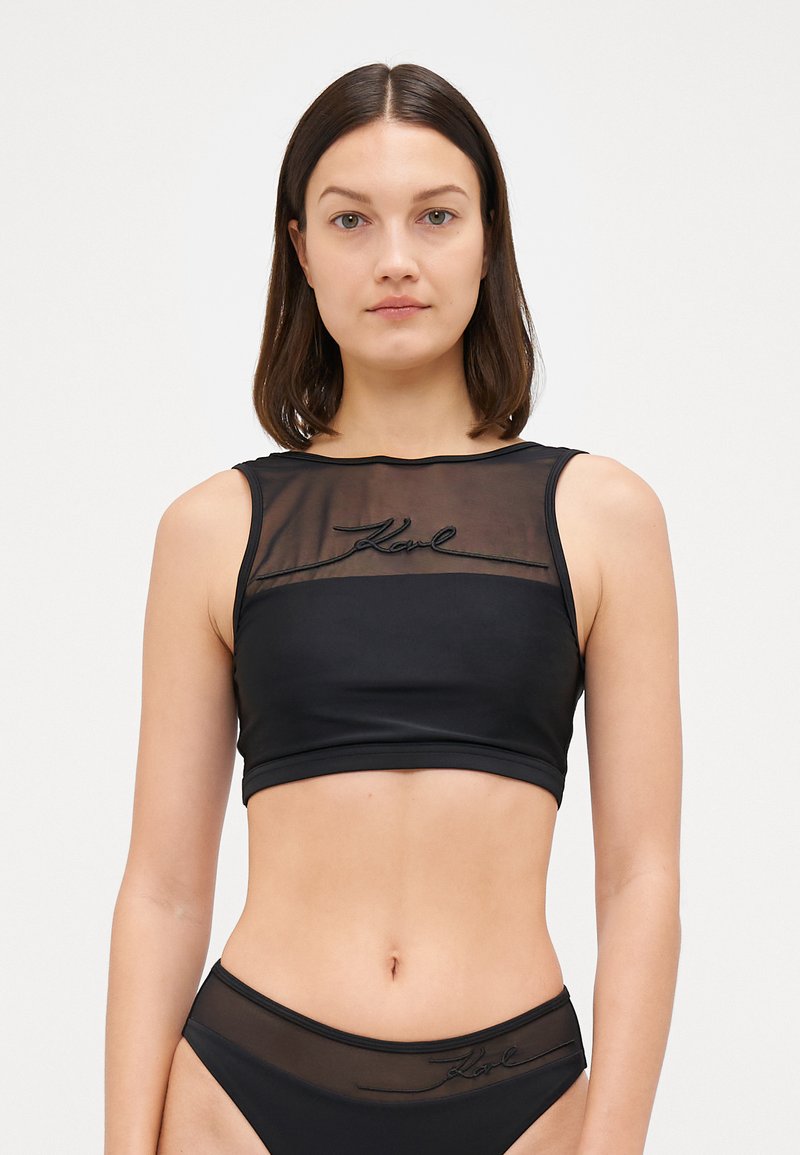 Svart crop top med en transparent mesh front, brodert logo og en figursydd design. Materialet har en glatt tekstur med en cropped cut.