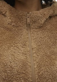 Krémová fleecová bunda s texturovaným, nadýchaným povrchem. Má přední zip a kapuci z měkkého materiálu, která poskytuje teplo a pohodlí.