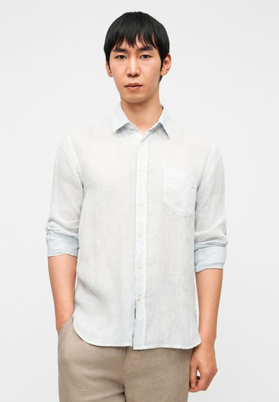 Ανοιχτό μπλε λινό πουκάμισο με κουμπιά, τσέπη στο στήθος, μανίκια που γυρίζουν και καμπυλωτό τελείωμα, συνδυασμένο με μπεζ παντελόνια.