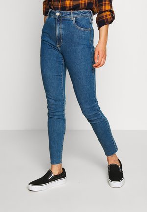Personne portant un jean skinny taille haute bleu avec des ourlets bruts, une chemise à carreaux marron et noire, et des baskets noires sans lacets avec des semelles blanches.