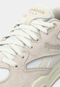 Detailný pohľad na krémové a biele tenisky Reebok s zamieňaným zvrškom z kože, sieťovanou podšívkou, bielymi šnúrkami a embosovaným logom na jazyku a boku.